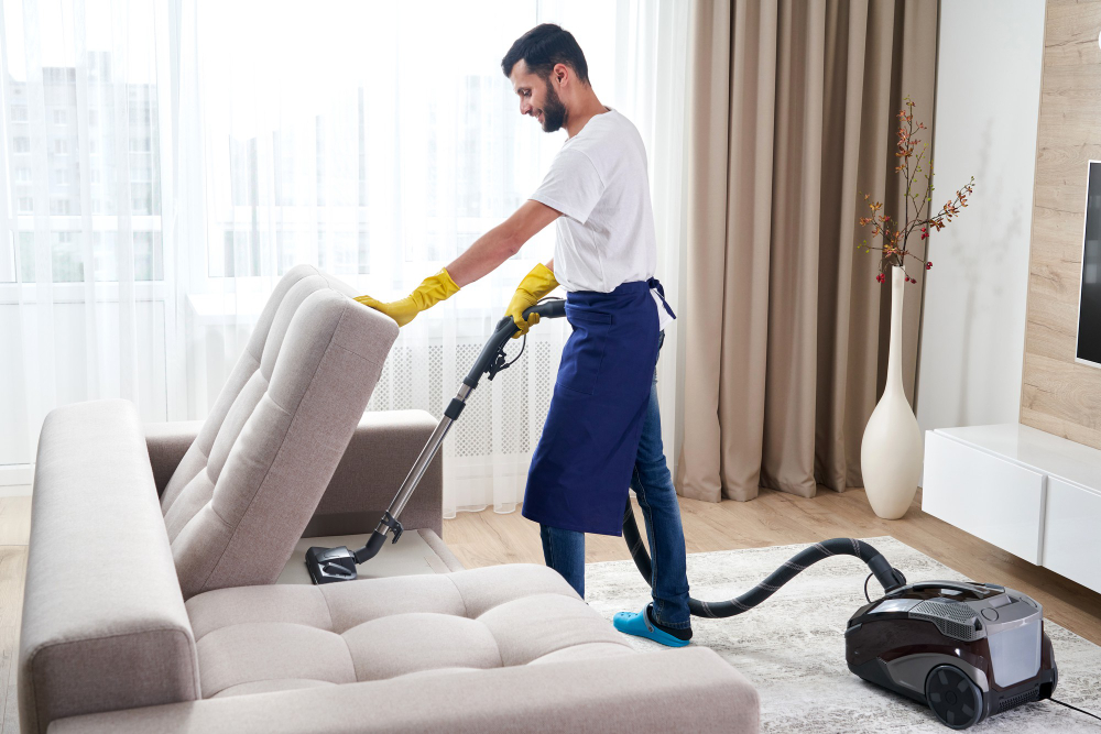 young-man-cleaning-sofa-with-vacuum-cleaner-leaving-room-home
